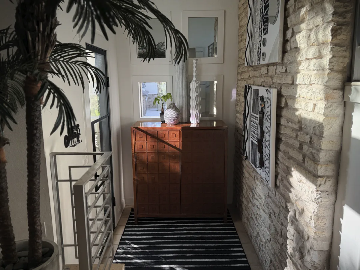 Late sun falls across a wooden cabinet with white vases, between a potted palm and a limestone wall hung with black and white art