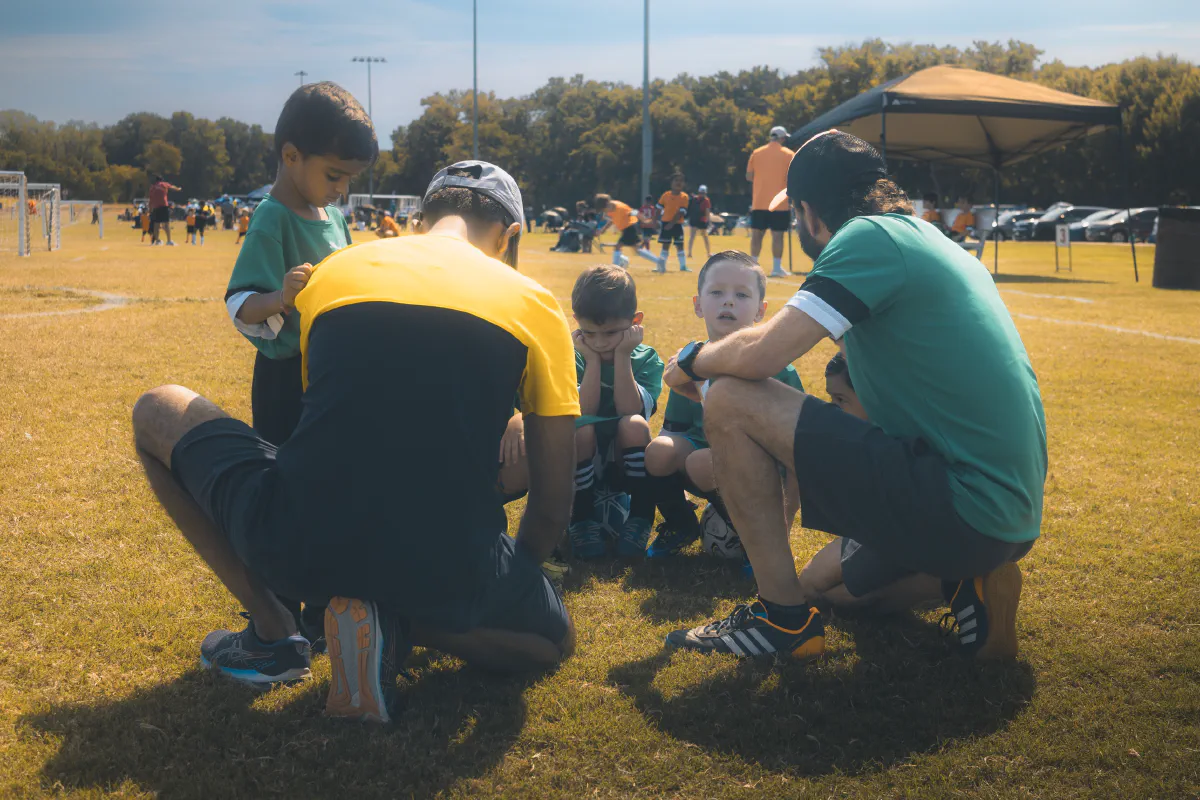Two coaches crouch in a huddle with young players in green jerseys on a sunny soccer field