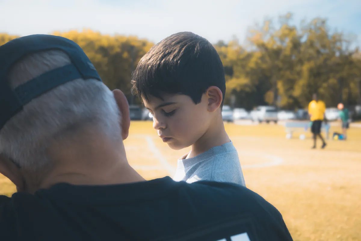 A boy looks down over the shoulder of an older man in a backwards cap, with the field blurred behind them