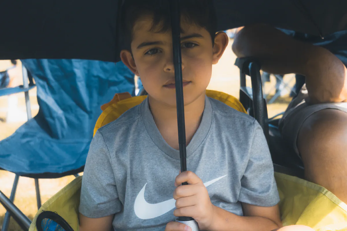 A boy in a gray shirt sits in a camp chair under an umbrella, its handle splitting his face down the middle