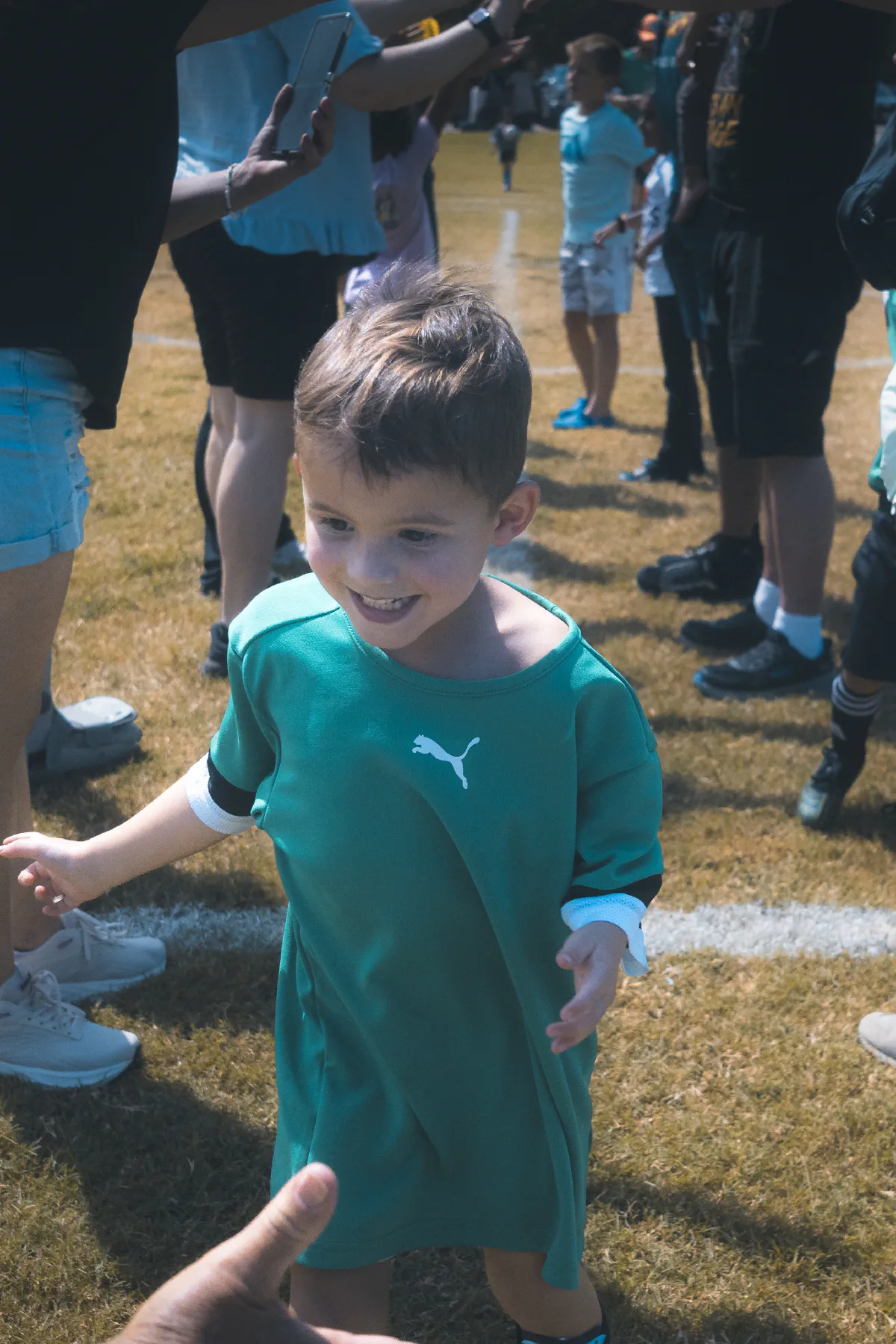 A grinning toddler in an oversized green jersey runs through a tunnel of parents' arms